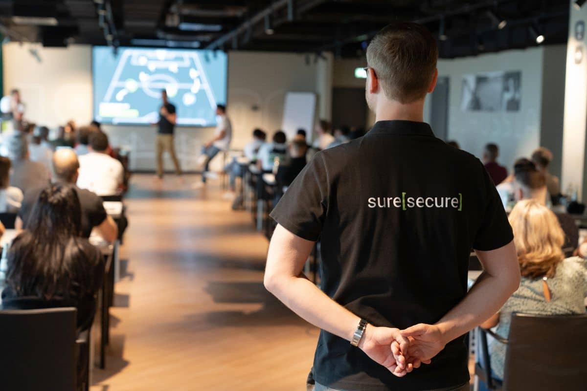 Ein Saal voller Menschen auf einer Konferenz und im Fokus sieht man einen Mann von hinten mit dem Logo der suresecure auf dem T-Shirt.